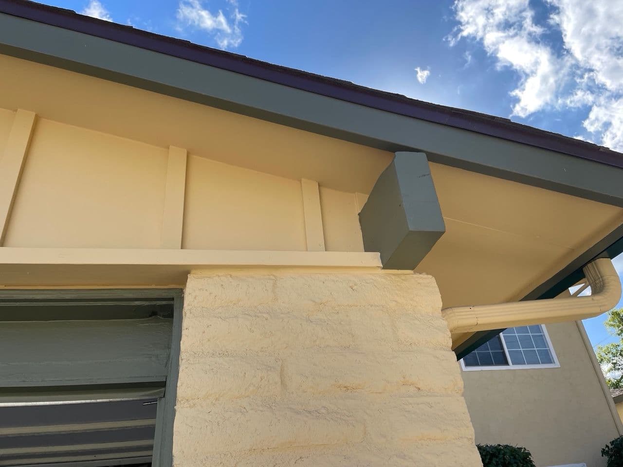 Close-up of a house corner with beige stucco, a green roof overhang, and clear blue sky.